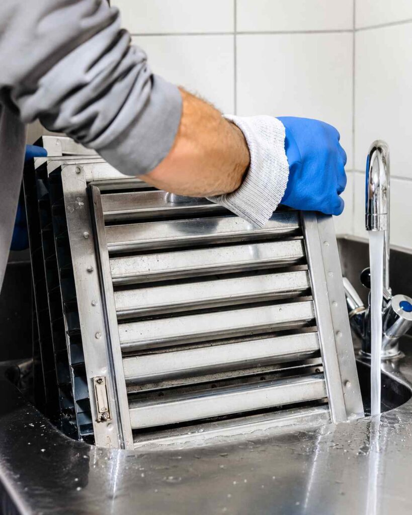 Technicien portant des gants rinçant un filtre de hotte professionnelle en inox dans un évier avant son dégraissage complet, lors d'une intervention de nettoyage Monaclean.